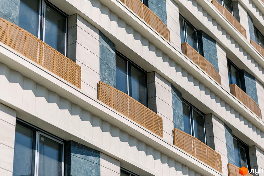 Фасад Житлового комплексу Einstein Concept House зі світлими рельєфними панелями, синіми вставками, великими вікнами та золотистими перфорованими огорожами.