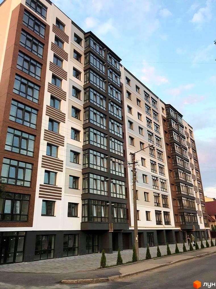 Фасад новобудови з коричневими та білими елементами, великими вікнами та заскленими балконами. Поруч тротуар з бруківки та зелені насадження.