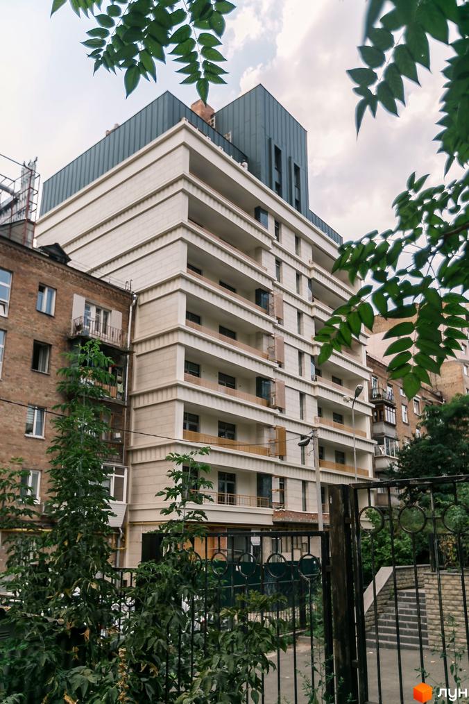 Новобудова Einstein Concept House має світлий фасад з балконами та темну верхню частину. Перед будинком видно металеву огорожу та зелені насадження.