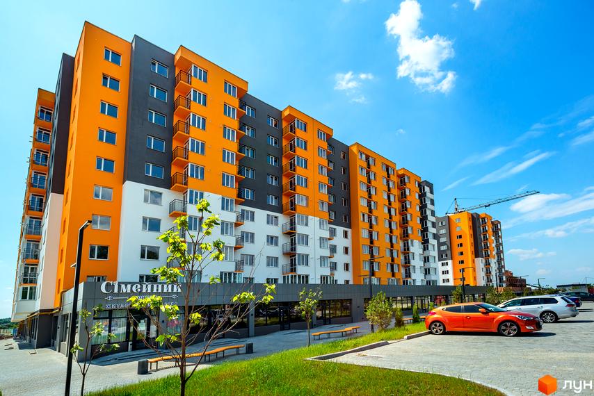 Житловий комплекс Сімейний COMFORT з яскравим фасадом з помаранчевих та сірих секцій. На прибудинковій території розташовані парковка та зелені насадження.