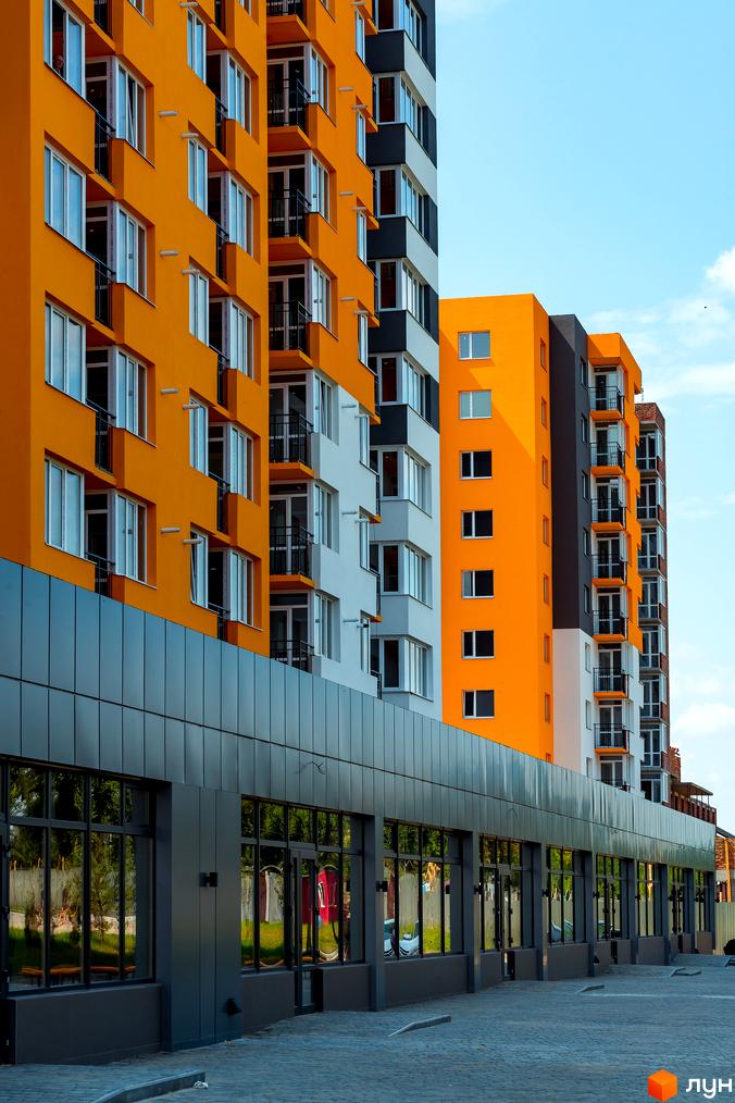 Новобудова Сімейний COMFORT з яскравим жовтим фасадом та великими вікнами. Поруч розташована комерційна зона з панорамним склінням та бруківкою.