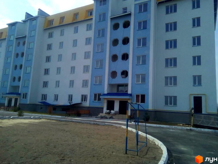 Зовнішній вигляд новобудови на вулиці Грушевського, 108а, з блакитними та білими фасадами та прибудинковою територією з піщаною зоною та дитячим майданчиком.