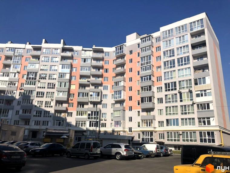 Зовнішній вигляд багатоквартирного будинку з сірими та персиковими фасадами, багато вікон та балконів. Біля будинку розташована парковка з автомобілями.