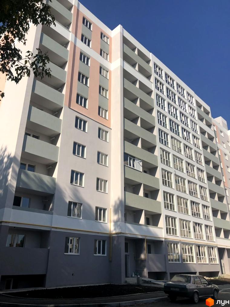 Зовнішній вигляд новобудови на вул. Жабинського, 2д, 2в, 2е. Будинок має сірі та рожеві елементи фасаду, балкони та багато вікон. Біля будинку розташована прибудинкова територія з дорогою та припаркованим автомобілем.