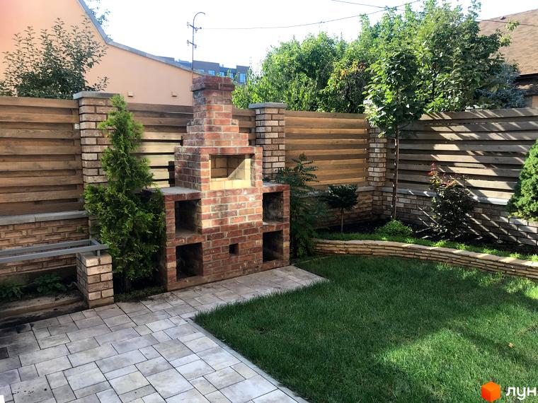 Таунхауси „На Достоєвського“: цегляний мангал та зелений двір з бруківкою. Затишна прибудинкова територія з деревами та кущами.
