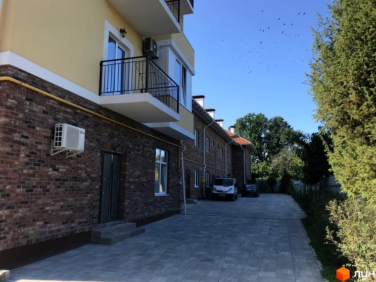 Зовнішній вигляд новобудови Таунхауси „На Достоєвського“ з цегляним фасадом та балконами. Прибудинкова територія з парковкою та деревами.