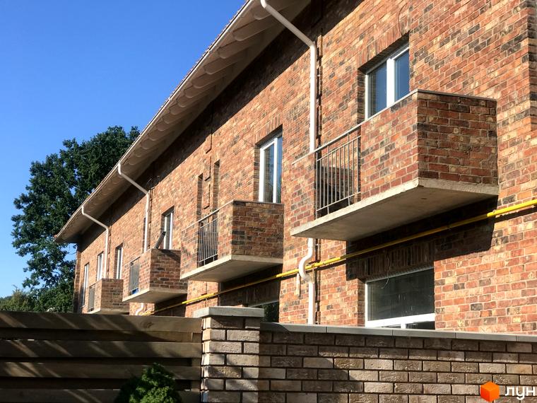 Зовнішній вигляд цегляних таунхаусів „На Достоєвського“ з балконами та вікнами на тлі блакитного неба та зелені. Прибудинкова територія з дерев'яним парканом.