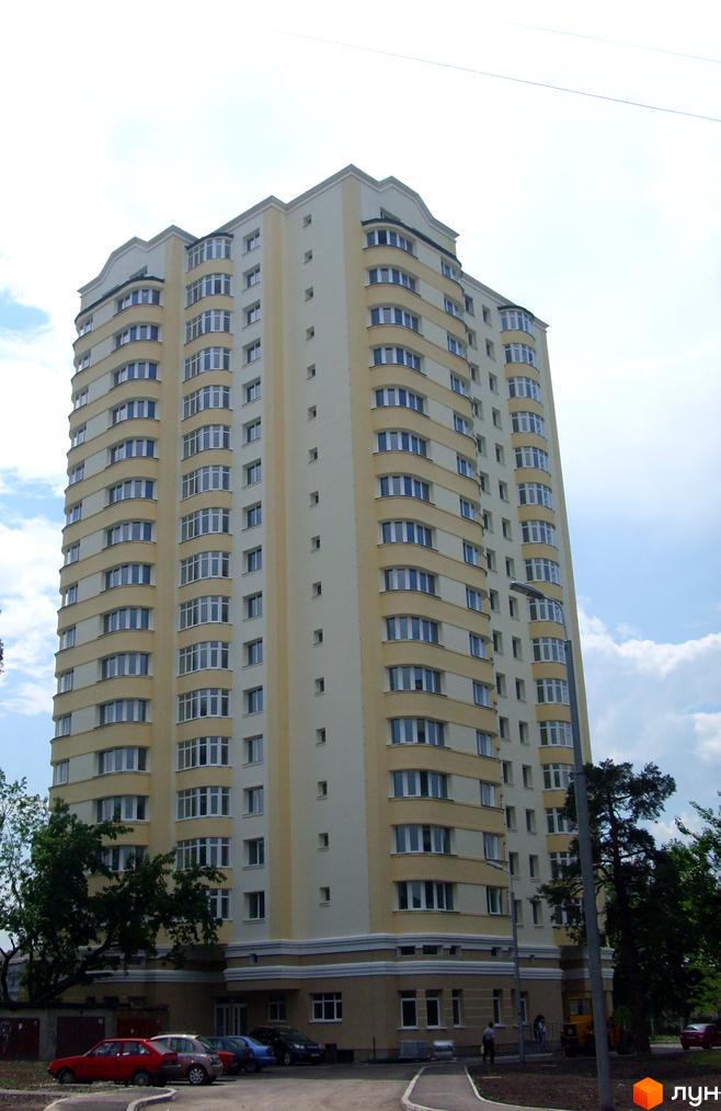 Багатоповерховий житловий будинок жовто-білого кольору з численними вікнами та округлими балконами. Перед будинком розташована парковка та зелені насадження.