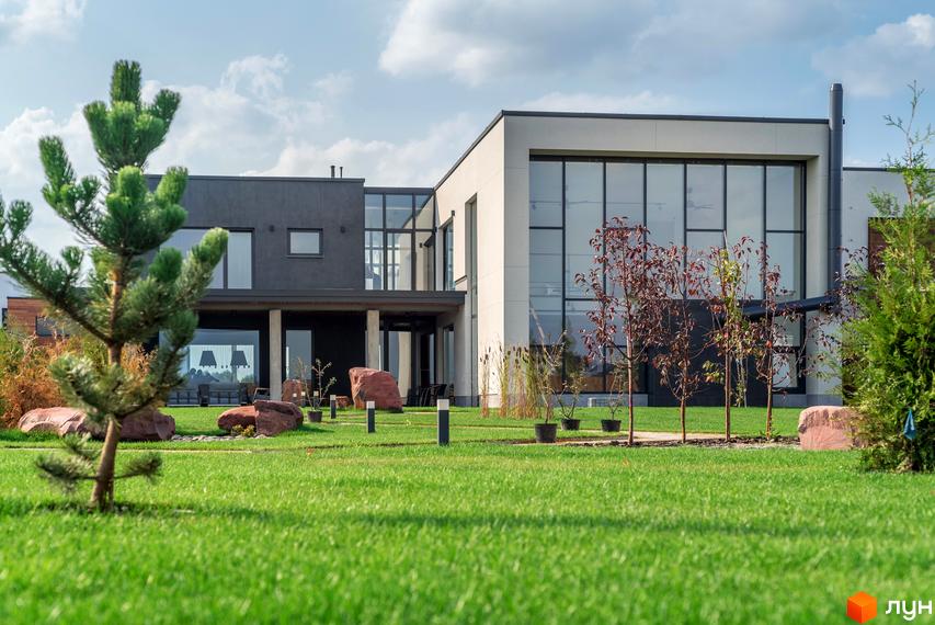 Котеджне містечко КМ На Щасливій. Сучасний двоповерховий будинок з великими вікнами та доглянутою зеленою прибудинковою територією з деревами та декоративними каменями.