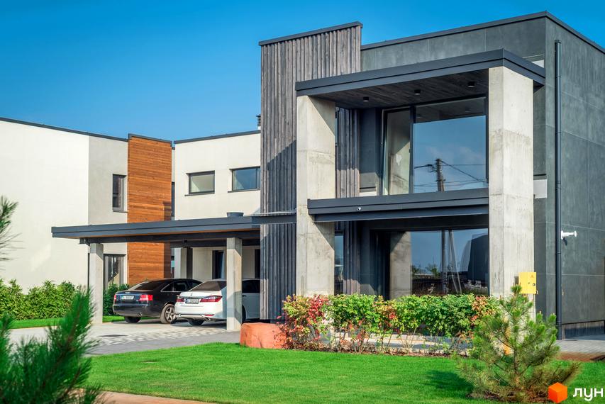 Котеджне містечко На Щасливій. Зовнішній вигляд будинку з бетону та дерева, з великими вікнами та навісом для авто. На прибудинковій території доглянутий газон та зелені насадження.