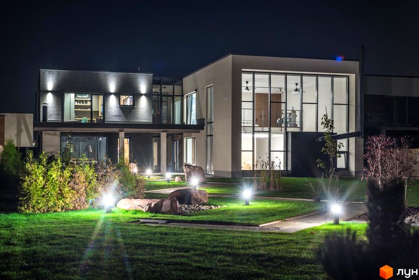 Котеджне містечко КМ На Щасливій. Нічна фотографія двоповерхового будинку з великими вікнами та підсвіченим фасадом. Прибудинкова територія з газоном, деревами та освітленням.