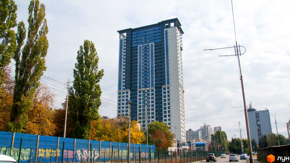 Висока новобудова ЖК Victory V з блакитними вікнами та білими стінами. Поруч ростуть високі дерева та пролягає дорога з автомобілями.