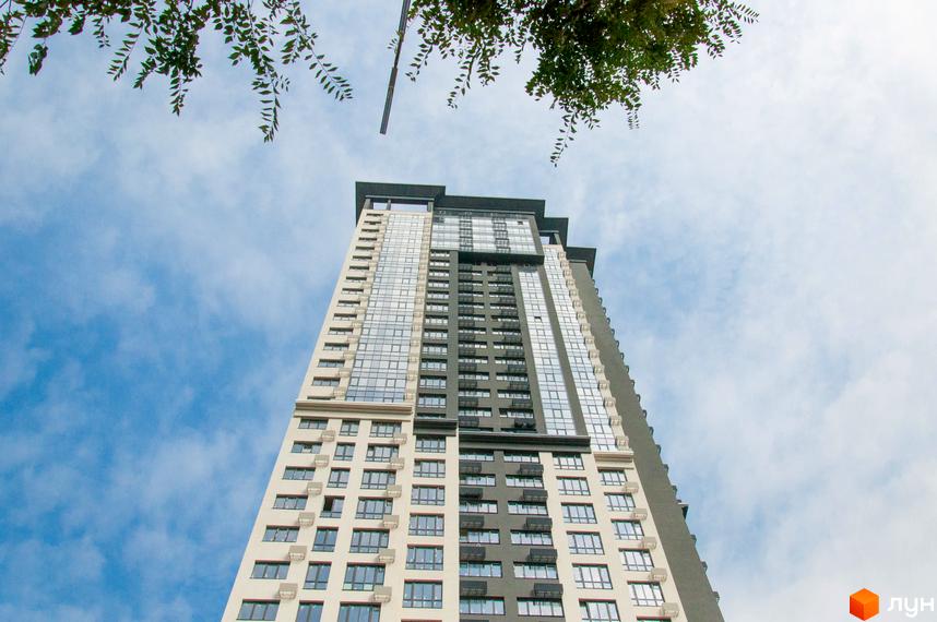 Новобудова Victory V: висотна будівля з контрастними світло-бежевими та темно-сірими фасадами, великою кількістю скляних вікон. Зверху видно зелене листя дерев на фоні хмарного неба.