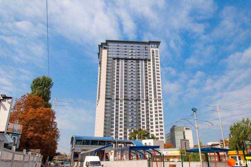 Високоповерховий житловий комплекс Victory V з сірими та білими фасадами, скляними балконами та темним дахом. Навколо видно дерева та міську інфраструктуру.