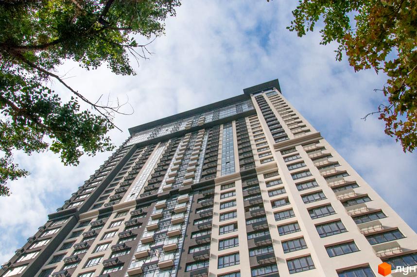 Зовнішній вигляд ЖК Victory V – висотної новобудови з балконами та панорамними вікнами на тлі хмарного неба. Навколо будівлі видно зелене листя дерев.