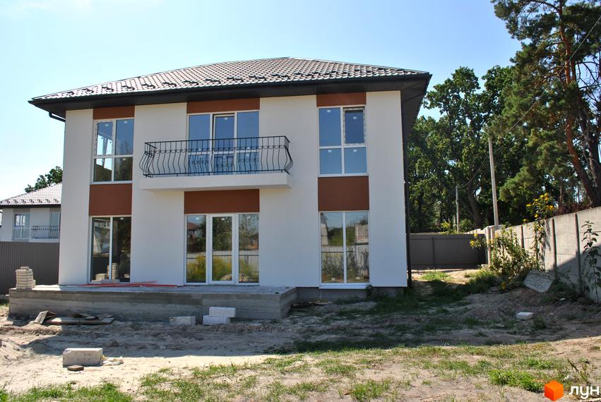 Будинок з білою фасадною штукатуркою та коричневими вставками, з балконом та великими вікнами. Територія навколо будинку в процесі будівництва, з будматеріалами та зеленими насадженнями.