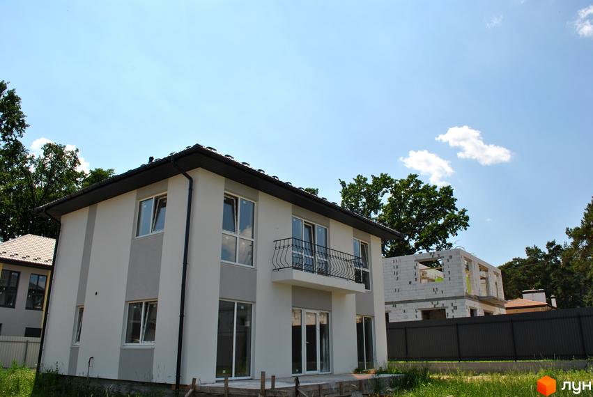 Зовнішній вигляд двоповерхового будинку з балконом та великими вікнами. На задньому плані видно будинок на стадії будівництва та зелені дерева.