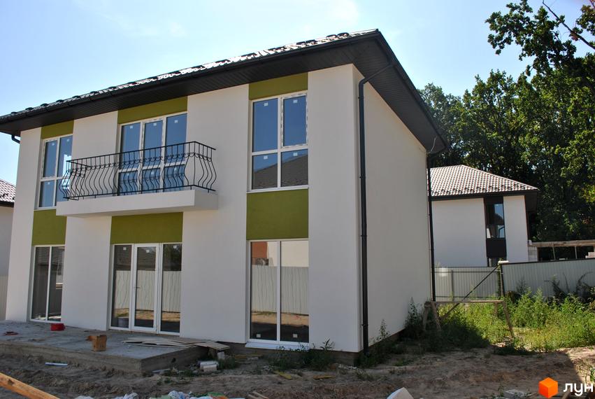 Зовнішній вигляд новобудови з білим фасадом та зеленими вставками, балконом та великими вікнами. Навколо будинку видно прибудинкову територію з деревами та будівельними матеріалами.