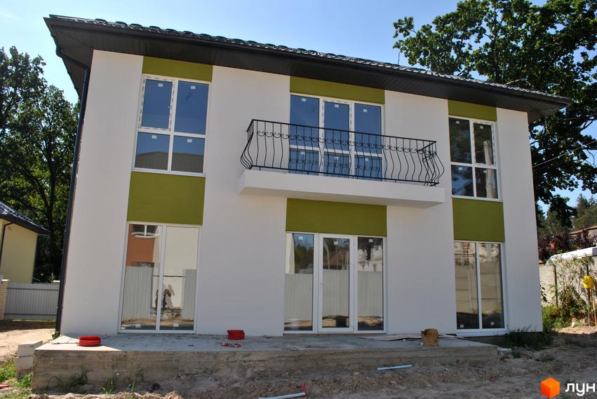 Зовнішній вигляд двоповерхового будинку з зеленими вставками та балконом. Навколо будинку видно будівельні роботи та дерева.