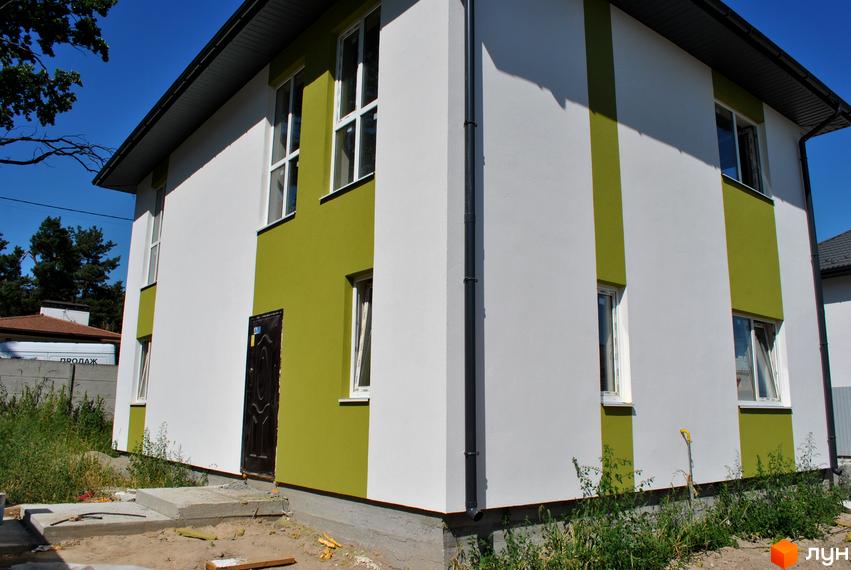 Зовнішній вигляд двоповерхового будинку з біло-зеленим фасадом та вікнами. Навколо будинку видно зелені насадження та будівельні матеріали.