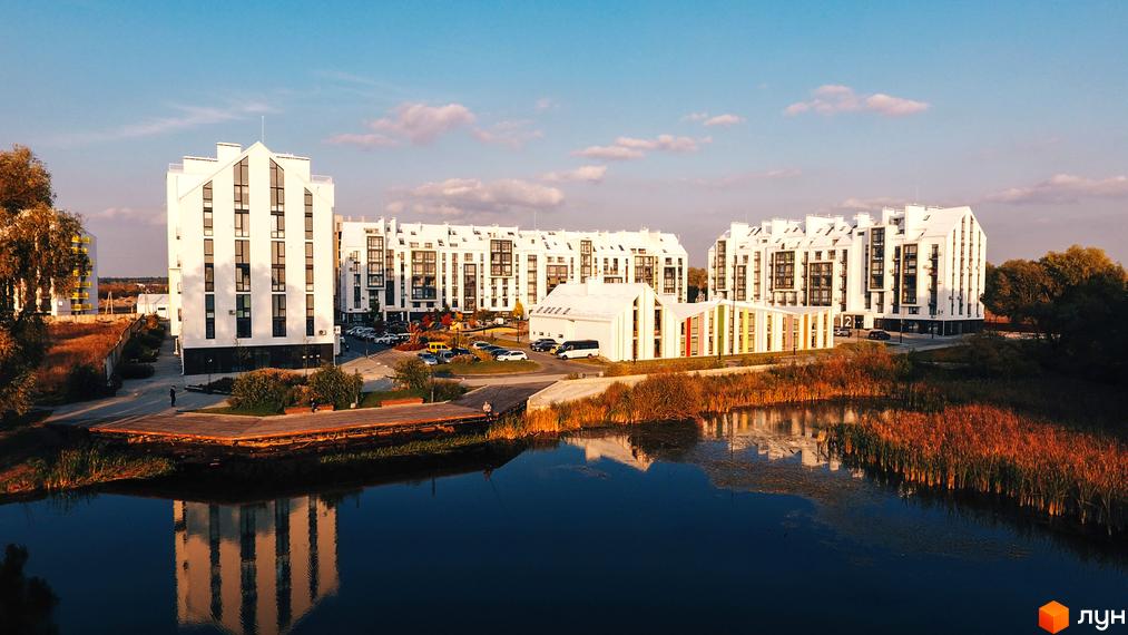 Вигляд на Житловий комплекс City Lake з білими багатоповерховими будинками та озером. На передньому плані очерет та дерев'яний пірс.