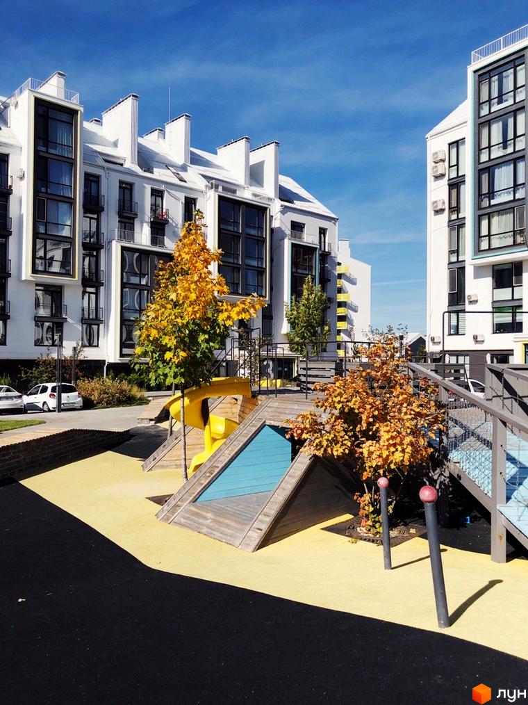 Житловий комплекс City Lake: білі багатоповерхові будинки з великими вікнами та балконами. На передньому плані дитячий майданчик з жовтим покриттям та осінніми деревами.