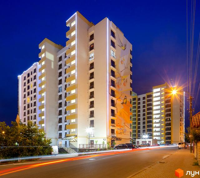 Житловий комплекс Lystopad у вечірній час. Фасади будівель прикрашені муралами та підсвічуванням. Повз новобудови проходить дорога з автомобільним рухом.