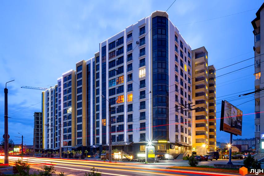 Житловий комплекс Lystopad – сучасна новобудова з яскравою архітектурою та підсвіткою фасадів. Комплекс розташований біля дороги з вечірніми слідами від автомобілів.