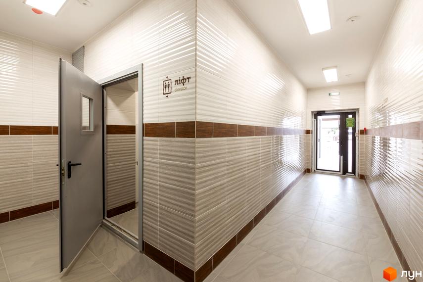 Коридор з ліфтом у новобудові ЖК Lystopad. Відкриті двері ліфта та вхідні двері до будинку. Стіни оздоблені плиткою.