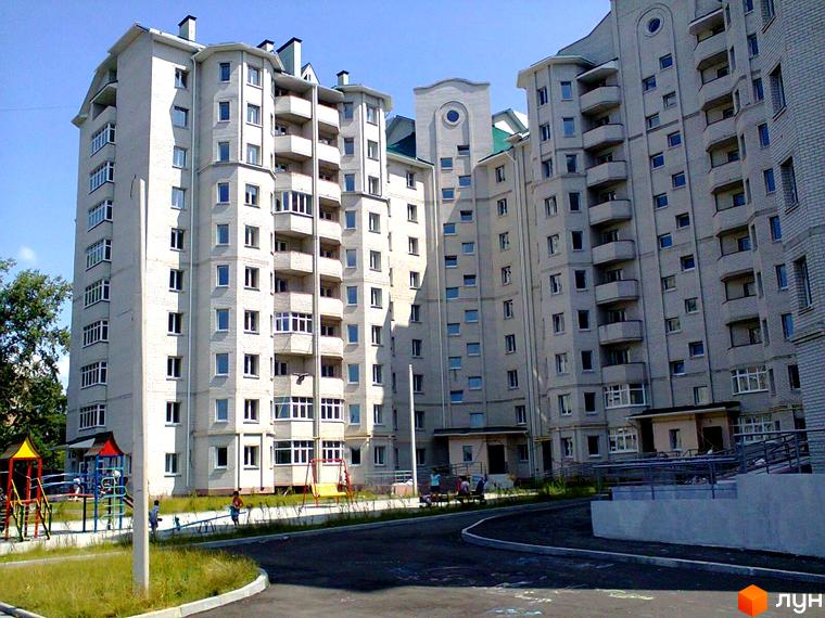 Багатоповерховий будинок з балконами та вікнами. На території є дитячий майданчик та асфальтована дорога.