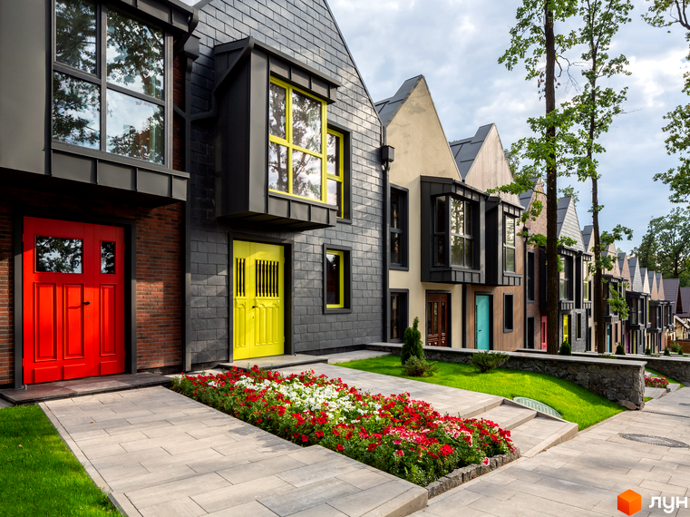 Зображення фасаду будинку в Містечку котеджів Oak Grove Town. Будинки з сірим дахом та яскравими дверима, перед будинками клумби з квітами та доріжки.