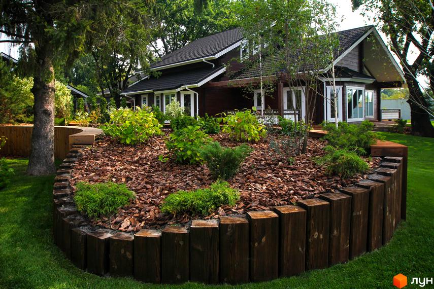 Котеджне містечко Riverside Family Club: двоповерховий будинок з темними стінами та білими вікнами, оточений доглянутим ландшафтом з газоном та декоративними рослинами.