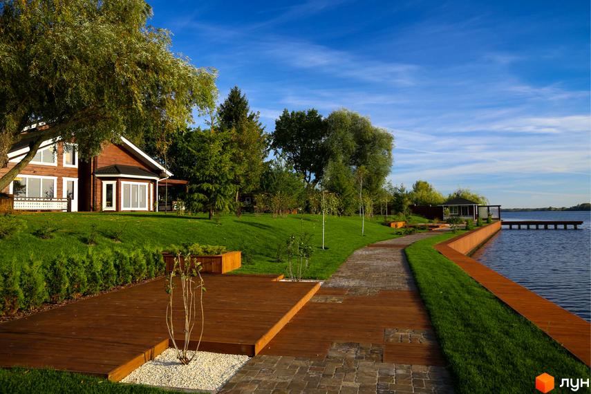 Котеджне містечко Riverside Family Club: двоповерховий дерев'яний будинок з великими вікнами, розташований на доглянутій зеленій ділянці з дерев'яною терасою біля озера.