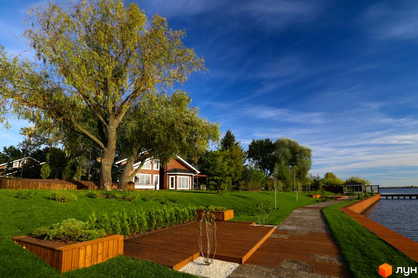 Котеджне містечко Riverside Family Club: двоповерховий дерев'яний будинок з великими вікнами на березі водойми. Доглянута прибудинкова територія з газоном, деревами та дерев'яною терасою.