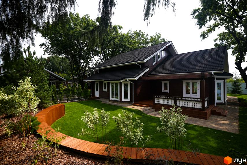 Котеджне містечко Riverside Family Club: дерев'яний будинок з білими вікнами та ганком. Навколо зелений газон та вигнута дерев'яна доріжка.