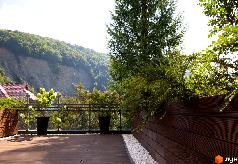 Житловий комплекс Крайобраз з терасою, що виходить на зелені схили гір. На терасі розташовані рослини в горщиках та дерев'яний настил.