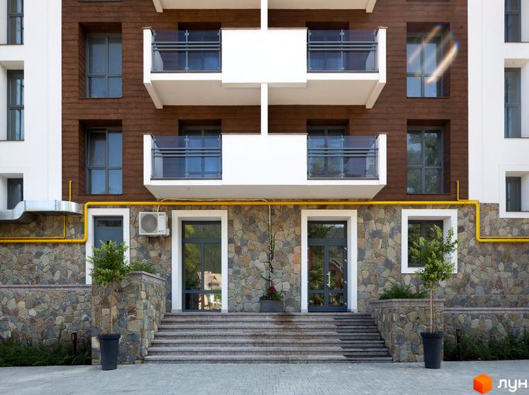 Фасад житлового комплексу Крайобраз з коричневою та білою обробкою, скляними балконами та кам'яною кладкою цоколя. Біля входу розташовані сходи та зелені насадження.