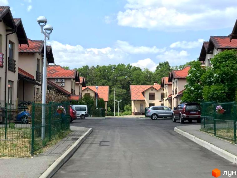 Котеджне містечко Садок Вишневий. Будинки з червоною черепицею та світлими фасадами. На території є асфальтована дорога, зелені насадження та припарковані автомобілі.