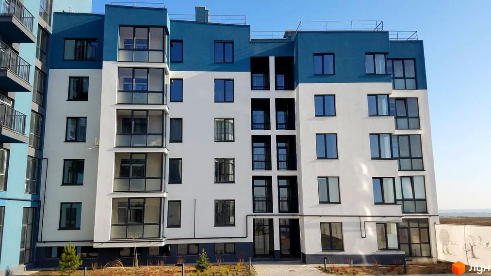 Новобудова ЖК Басів схил з біло-синім фасадом та великою кількістю скляних балконів. Навколо будинку облаштована прибудинкова територія з молодими деревами та газоном.