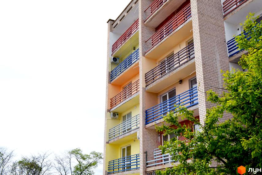 Апарт-комплекс Petrovets має яскраві фасади з балконами, обладнаними синіми та червоними поручнями. Навколо будівлі ростуть зелені дерева.