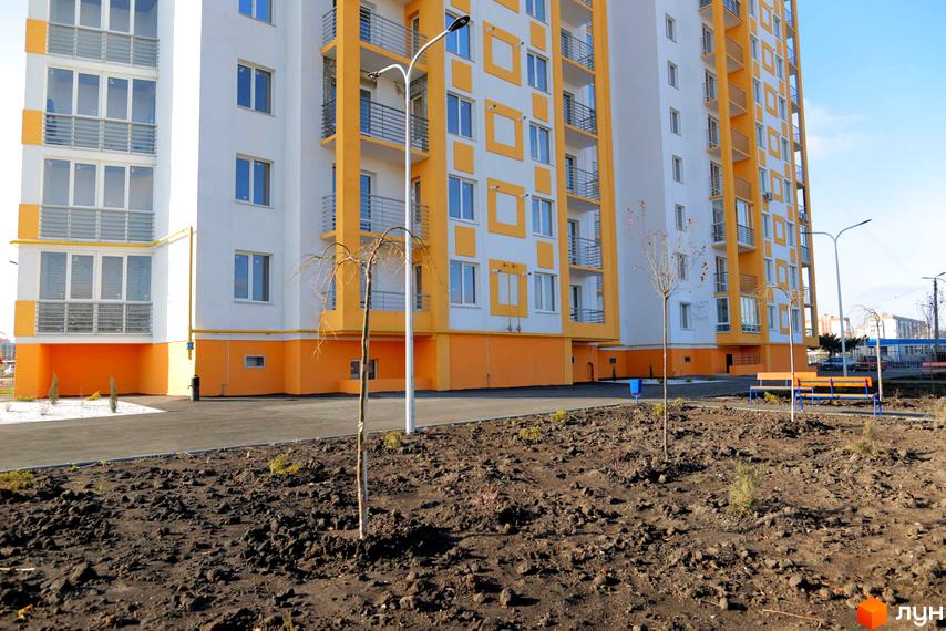 Житловий комплекс Апельсин. Зовнішній вигляд новобудови з жовто-білим фасадом та балконами. Поруч облаштована прибудинкова територія з лавками та деревами.
