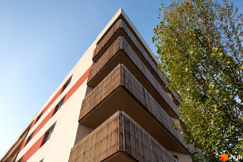 Зовнішній вигляд житлового комплексу Козацький Гай з балконами, оздобленими дерев'яними рейками, та червоною цегляною кладкою. Навколо будівлі зелене дерево під яскравим небом.