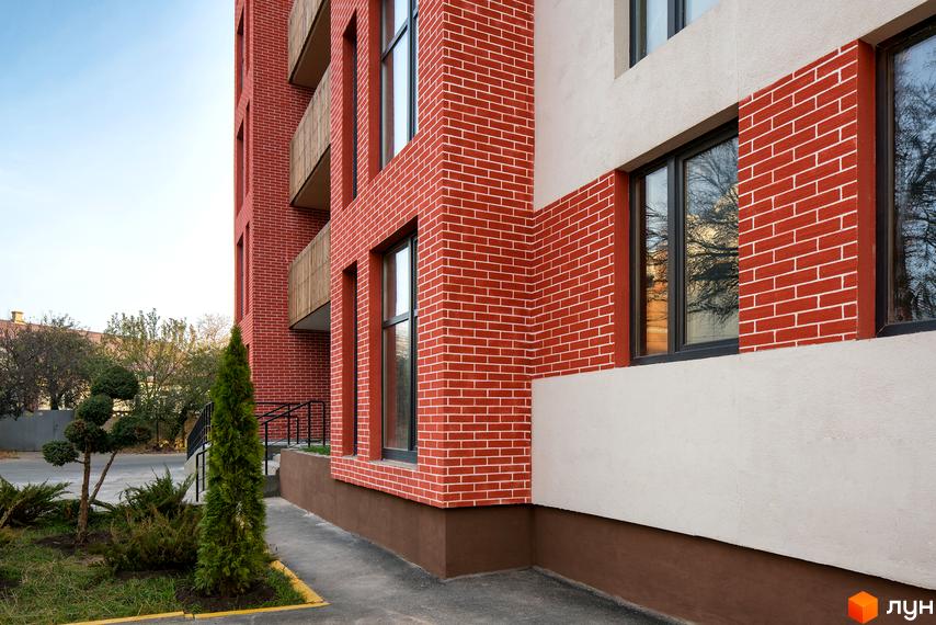 Зовнішній вигляд житлового комплексу Козацький Гай з червоної цегли та світлої штукатурки. Поруч доглянута прибудинкова територія з деревами та газоном.
