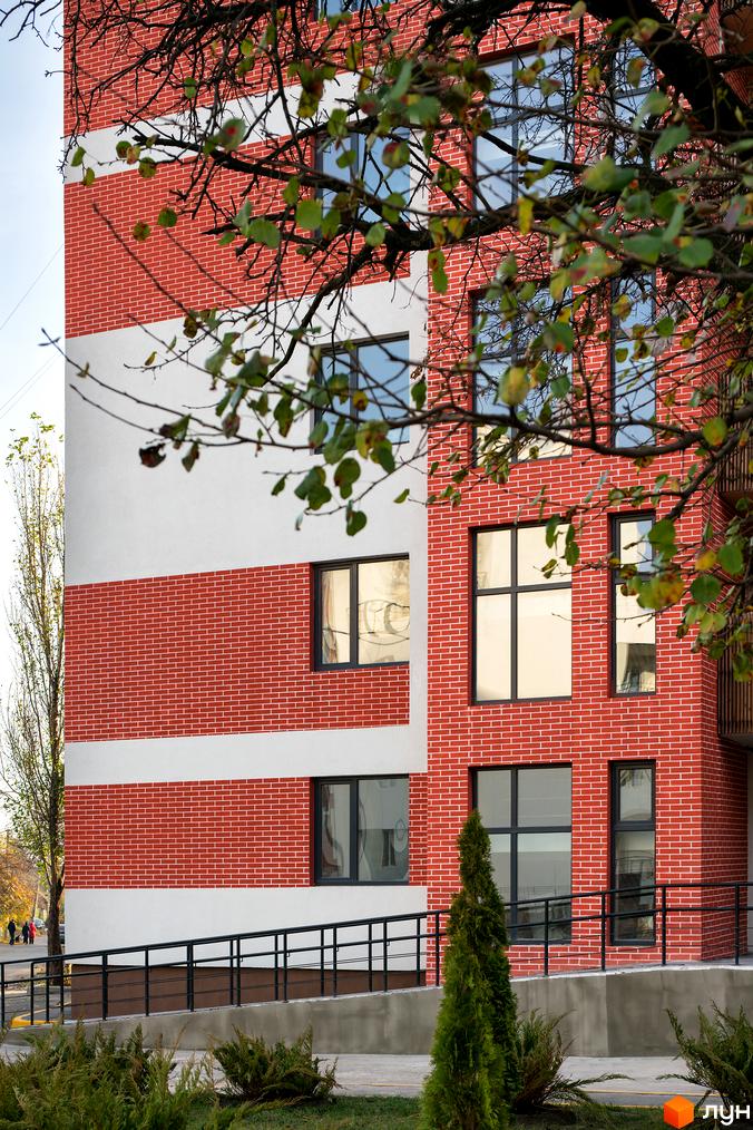 Фасад житлового комплексу Козацький Гай з червоної цегли та білої штукатурки, вікна з темними рамами. Поруч розташована прибудинкова територія з деревами та кущами.