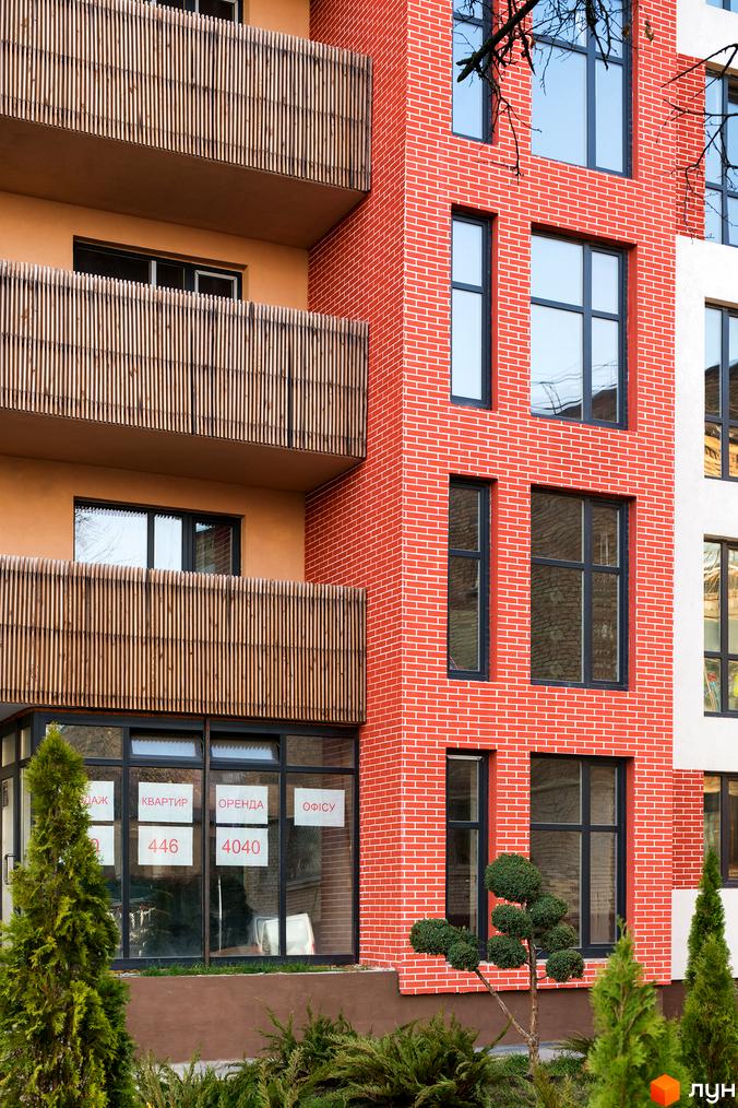Фасад новобудови ЖК Козацький Гай з червоної цегли та балконами з дерев'яними рейками. Біля будинку зелені дерева та кущі.