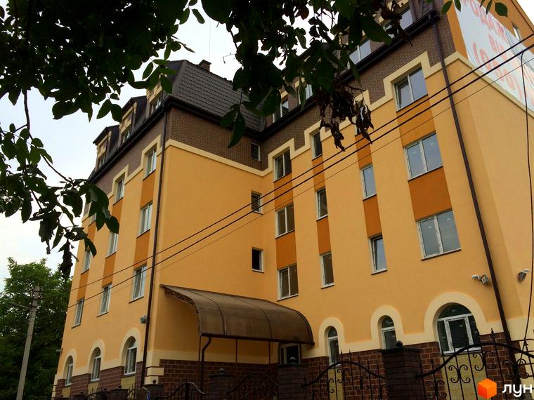 Фасад новобудови ЖК Еко Таун з жовтими та коричневими елементами, вікнами та дахом. На прибудинковій території видно паркан та зелені насадження.