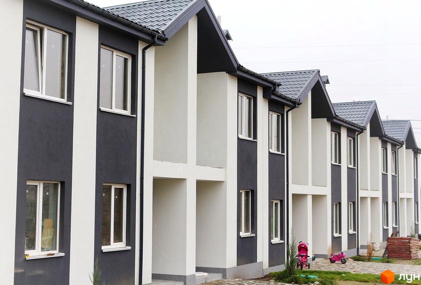 Зовнішній вигляд двоповерхових будинків з темним дахом та контрастним фасадом. Поруч дитячі візочки та будматеріали.