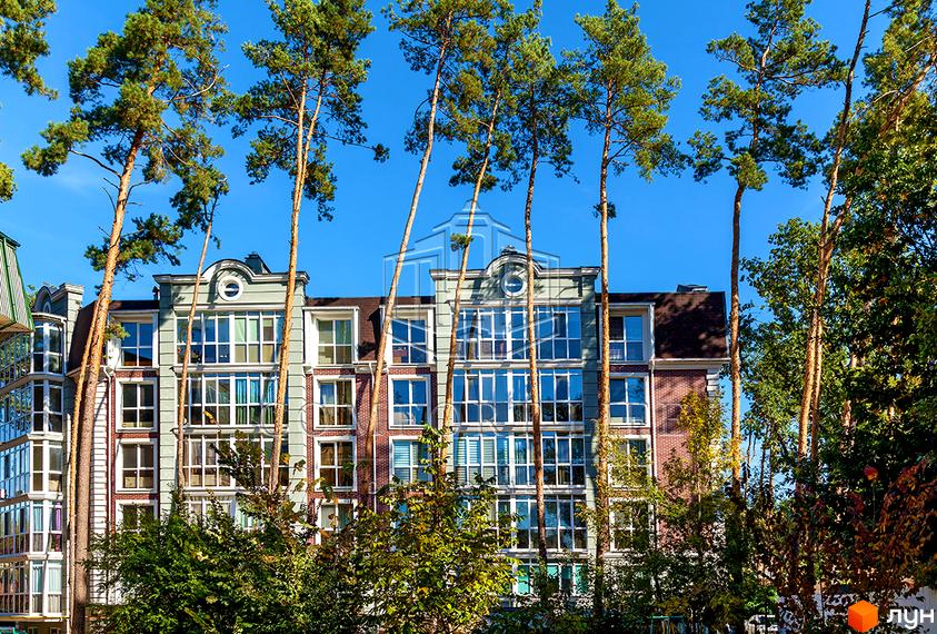 Зовнішній вигляд житлового комплексу Британіка з цегляними фасадами та великими вікнами, оточений високими соснами. Навколо комплексу багато зелені та дерев.