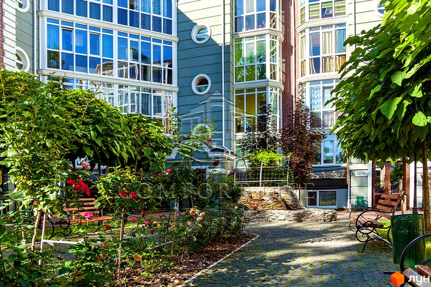 Зовнішній вигляд новобудови ЖК Британіка з великою кількістю вікон та балконів. Прибудинкова територія з зеленими насадженнями, квітами та місцями для відпочинку.