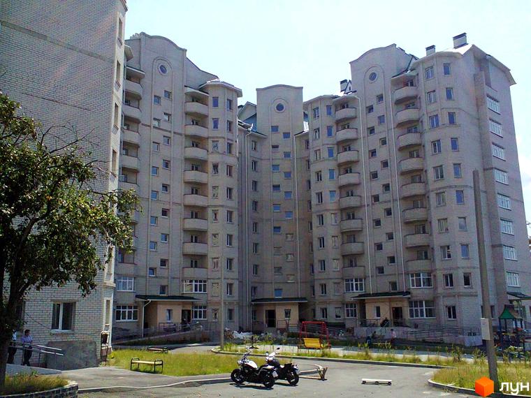 Багатоповерховий будинок з балконами та великими вікнами. На території комплексу є дитячий майданчик, лавки та місце для паркування мотоциклів.